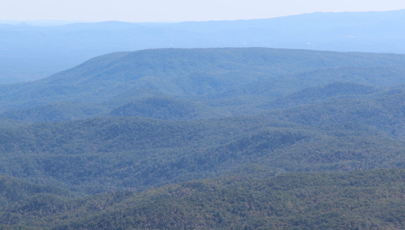 Long, flat-topped forested ridge of Brown Mountain under hazy blue sky, with layered blue mountains behind it.