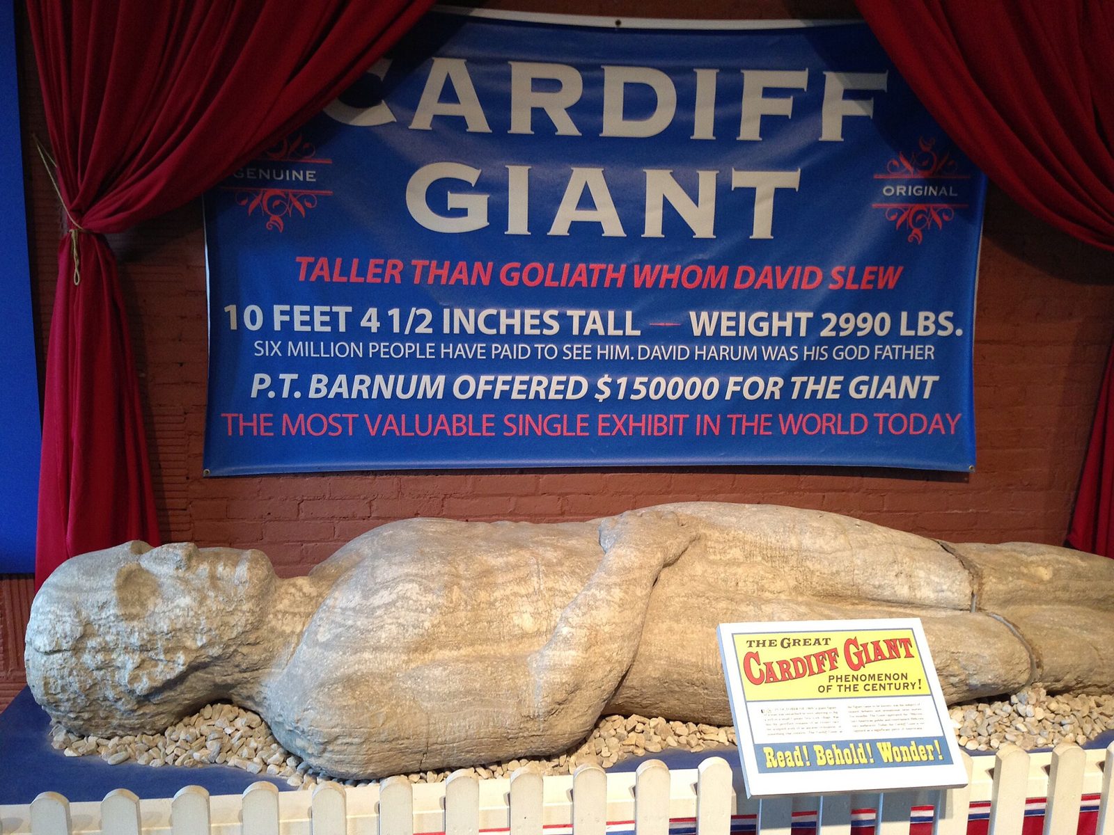 The pale, roughly carved stone figure of the Cardiff Giant lying in a gravel bed beneath a showman's banner that reads ‘Cardiff Giant, taller than Goliath whom David slew’.