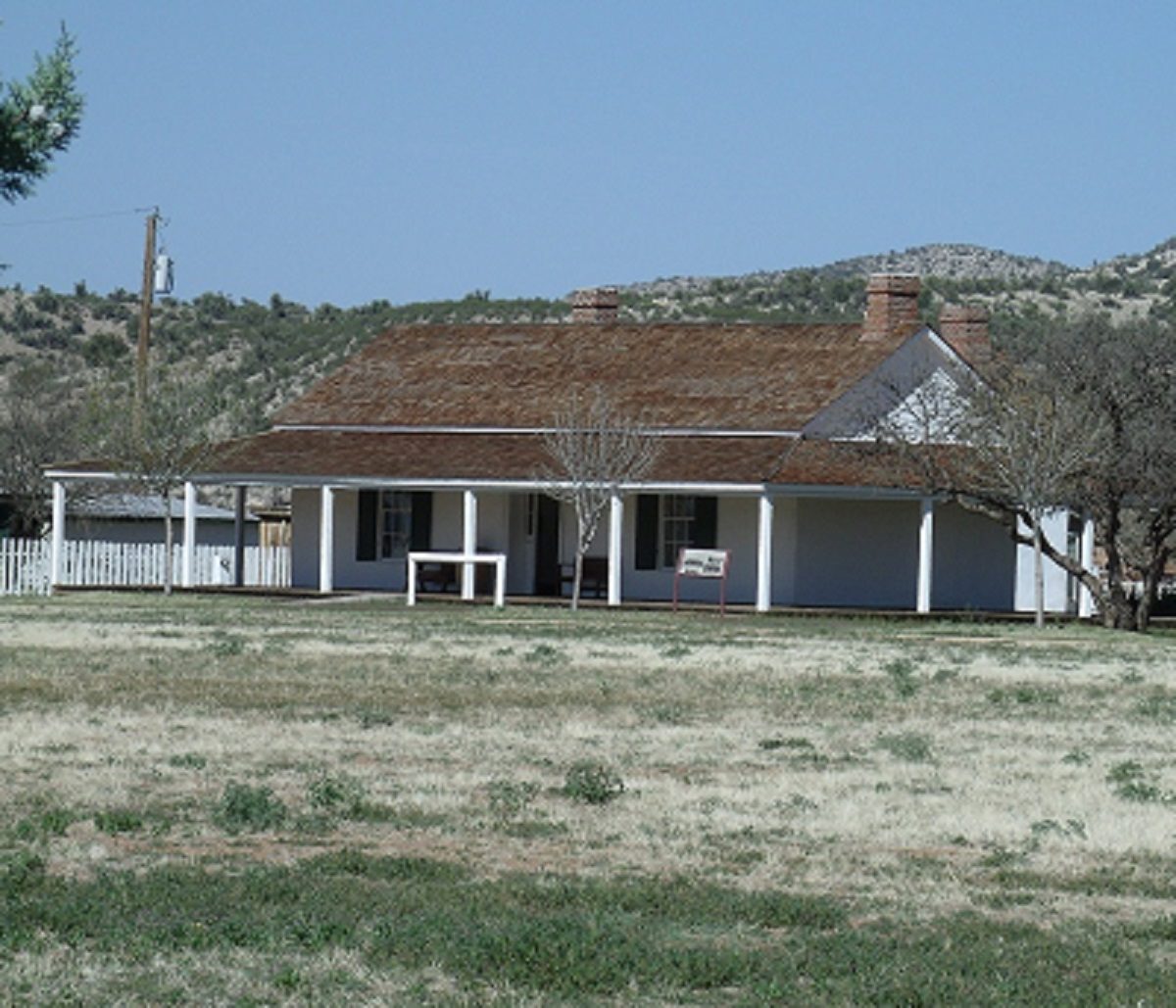 Single-story white frame house with a long front porch and brown shingled roof with two chimneys, set behind a grassy parade ground with desert hills behind.