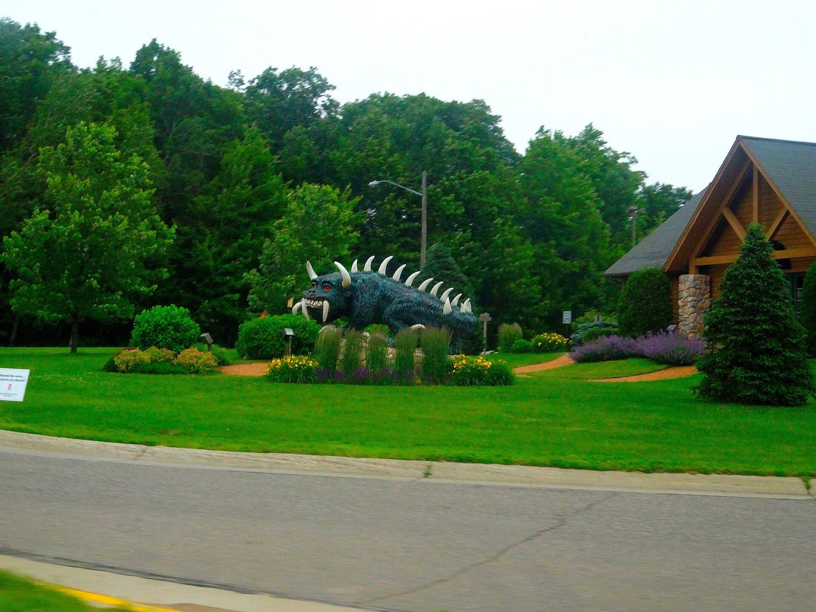 A large dark-green horned Hodag sculpture with white tusks and spines, set in a flower bed on a lawn beside a timber-framed visitor center.