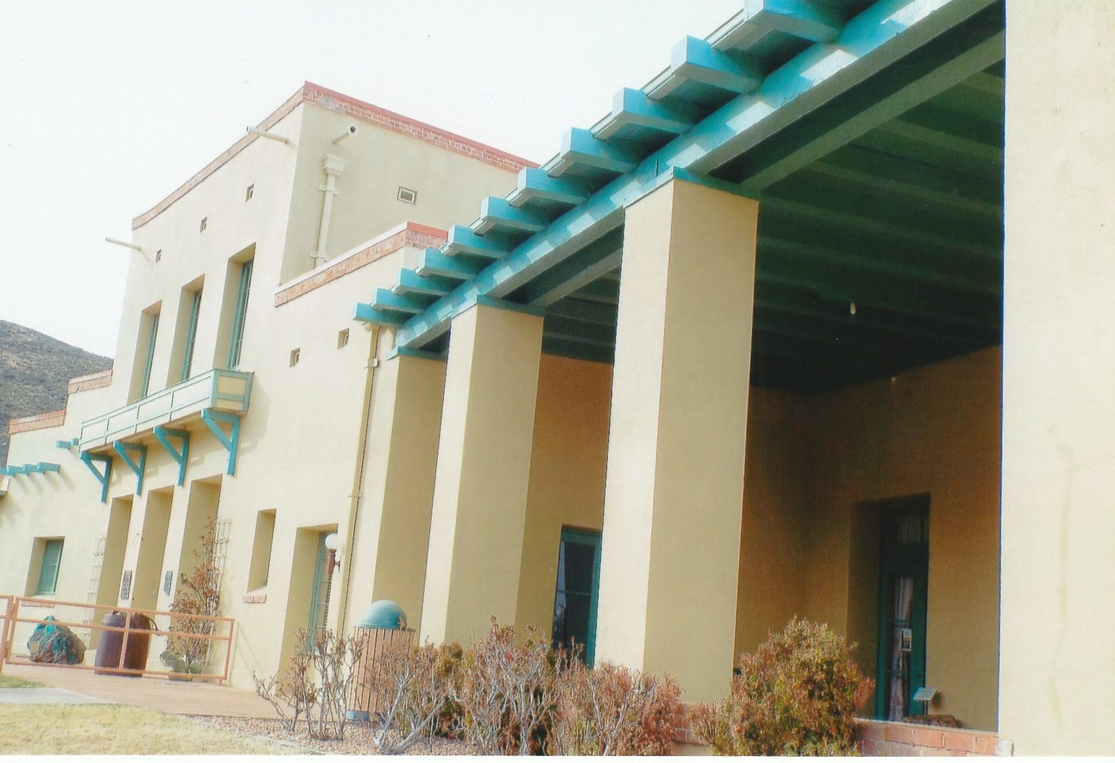 Cream-colored adobe-style two-story mansion with a turquoise-trimmed balcony and a deep columned porch, a rocky hillside behind it.