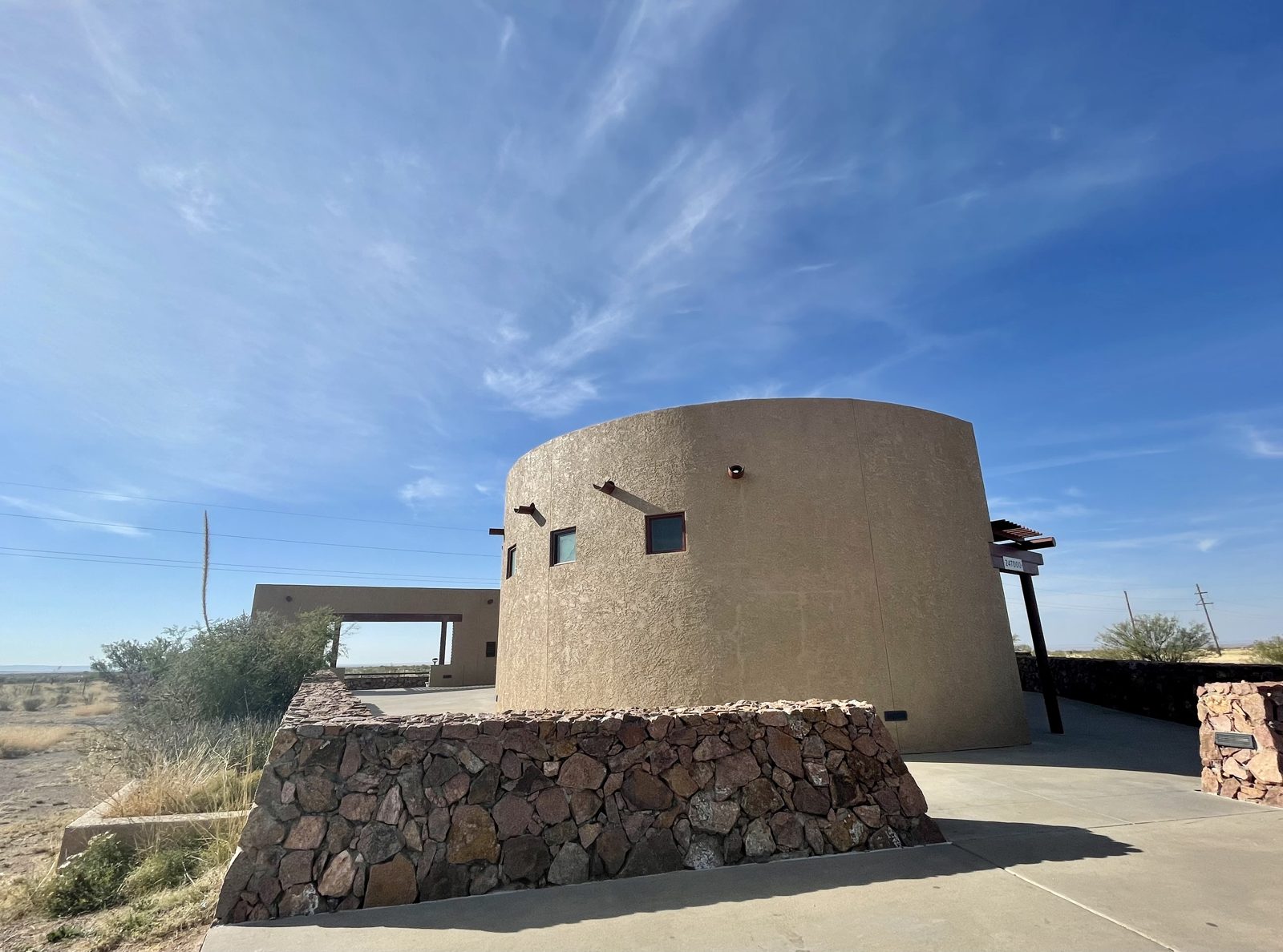 Round, tan stucco viewing building with small square windows and a stone wall in front, under a bright blue sky with wispy clouds in open desert grassland.