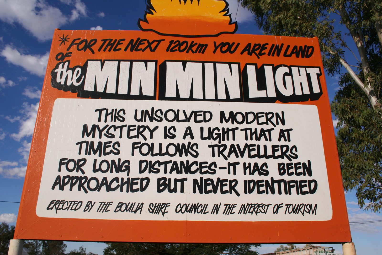 Bright orange roadside sign reading 'For the next 120 km you are in land of the Min Min Light. This unsolved modern mystery is a light that at times follows travellers for long distances. It has been approached but never identified. Erected by the Boulia Shire Council in the interest of tourism.'