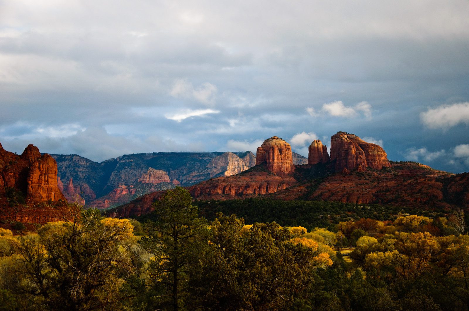 Red sandstone buttes and spires lit by late sun under dark storm clouds, above a band of green trees and yellow autumn foliage.