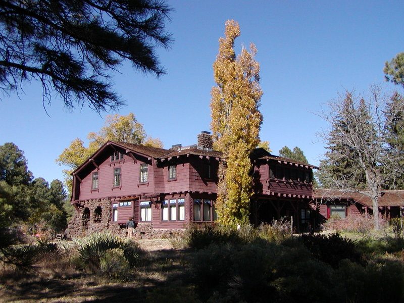 A large two-story dark-red shingled mansion with a stone lower wall, under a tall golden poplar and blue sky, surrounded by pines.