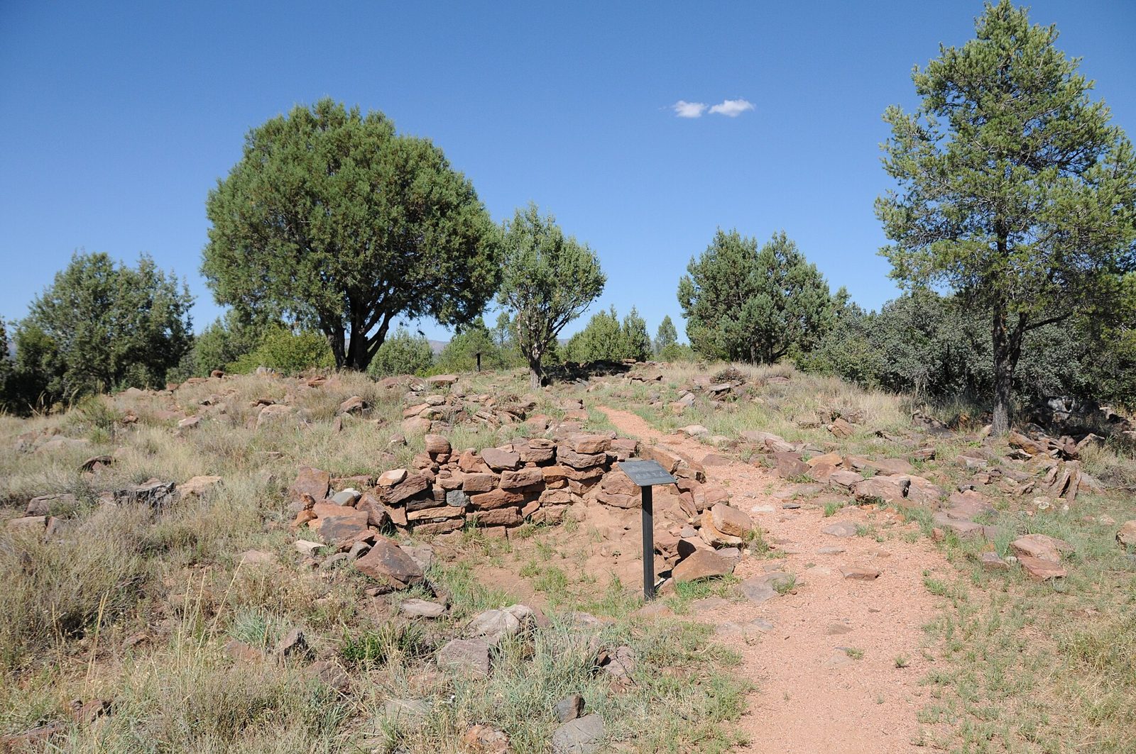 Low, partly collapsed walls of stacked reddish stone beside a dirt path and a small interpretive sign, among dry grass and juniper trees under a clear blue sky.