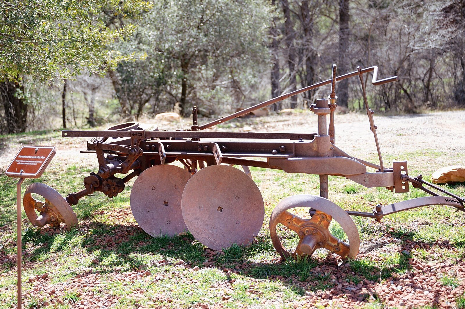 A rusted antique disc plow on iron wheels, set on grass beside a gravel path with a small interpretive sign and trees behind it.