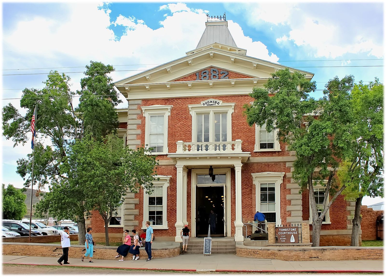 Red-brick two-story Victorian courthouse with white trim and a cupola, trees and visitors out front